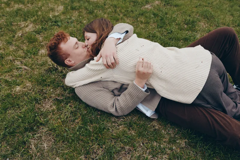 Young couple with red hair lies on grass in warm sweaters embracing each other tenderly enjoying a cozy lifestyle moment outdoors in autumn season.