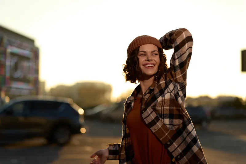 woman smile golden hour candid authenticity urban beanie plaid: joyful young woman laughing outdoors in a parking lot during warm sunset glow, emotional storytelling and mindful living vibe