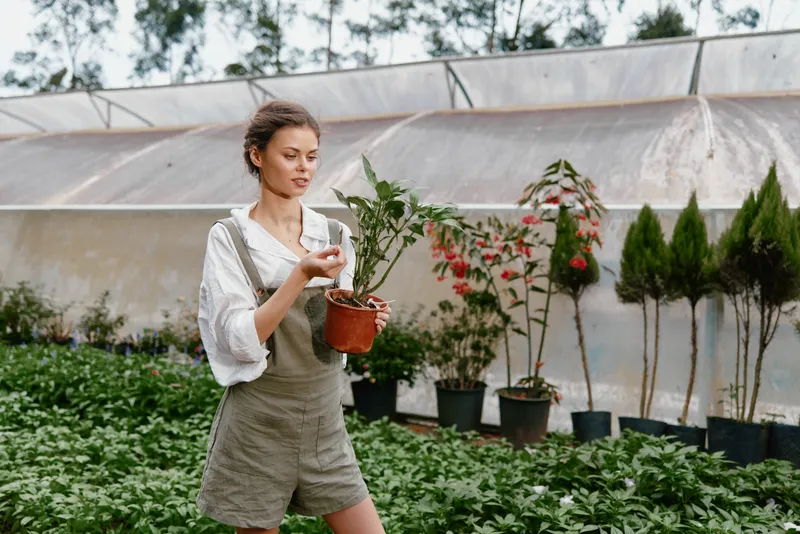 Young woman in casual overalls tending to plants in a greenhouse, smiling and enjoying gardening, surrounded by lush greenery and vibrant flowers