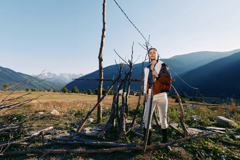 Woman portrait and traveler standing by rustic wooden fence in meadow below mountains, landscape and nature scene with clear sky, sunlight and autumn fashion jacket and boots.
