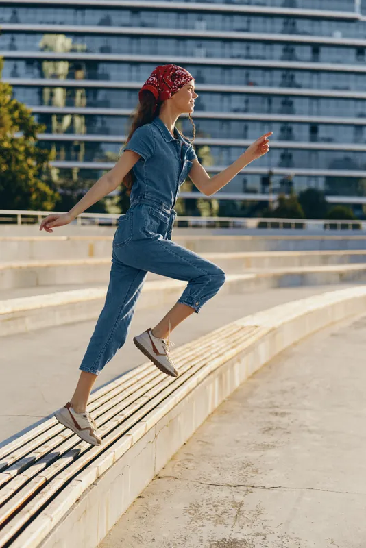 Lifestyle woman in modern boho-western denim street style jumps outdoors wearing casual shoes and red bandana, captured with warm film color tones in urban setting.