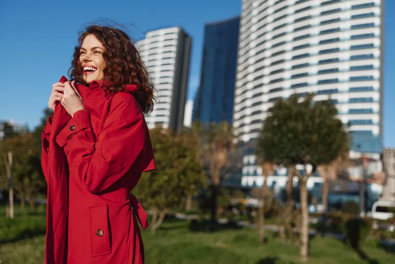 Young woman in a bright red coat smiles in a city park, sunlight on her curly hair with modern high rise buildings behind, in soft breeze and clear sky
