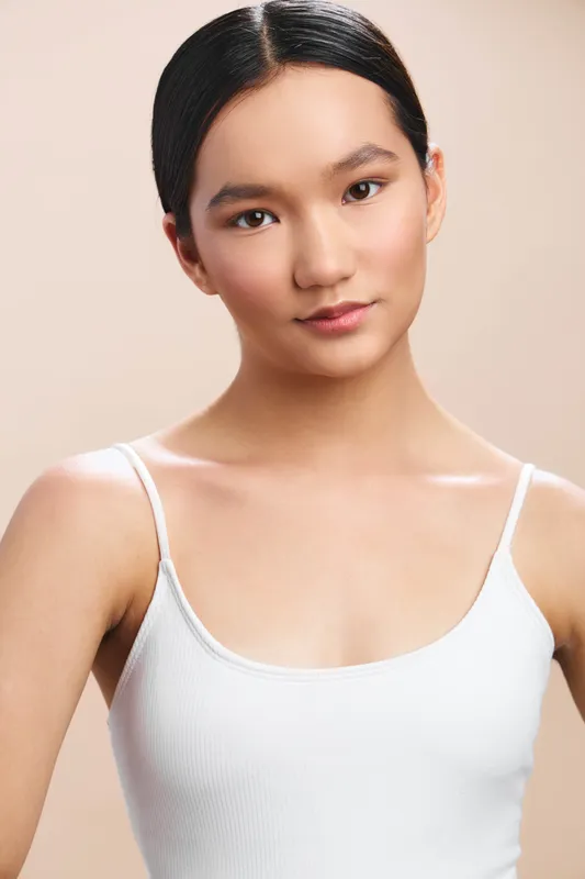 Asian woman gazes softly at the camera, wearing a white tank top against a neutral backdrop. A calm, confident portrait with clean lighting and minimal styling for versatile use.