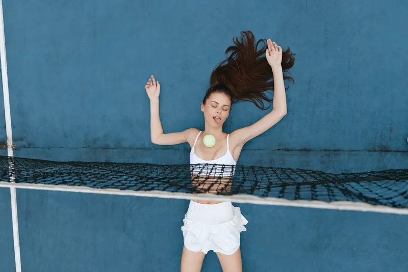 Young athletic woman enjoying summer leisure with a tennis ball, wearing a stylish white outfit against a vibrant blue background, conveying joy and energy