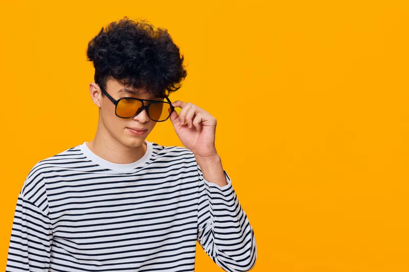 Young man wearing striped shirt and sunglasses posing confidently against vibrant orange background with curly hair and casual style.