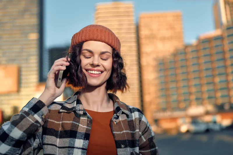 Woman smiling with eyes closed on phone in city at golden hour, candid lifestyle portrait in beanie and plaid jacket, authenticity and emotional storytelling, mindful living moment.