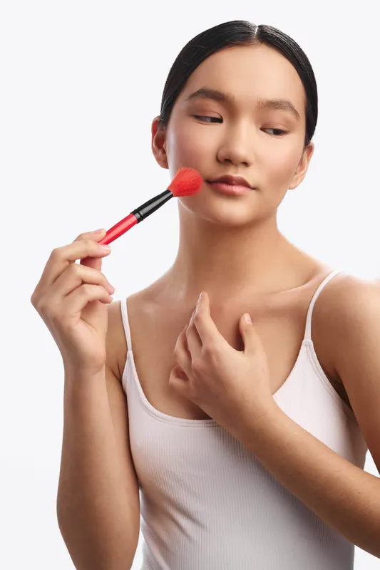 asian woman applies makeup with a soft smile in a clean studio, holding a brush to her cheek, her calm, confident expression conveying poise and self care.