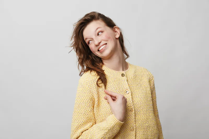 Smiling young woman with curly hair wearing yellow knitted sweater looks sideways with playful expression on light grey background. Concept of happiness, casual style, and positive mood.