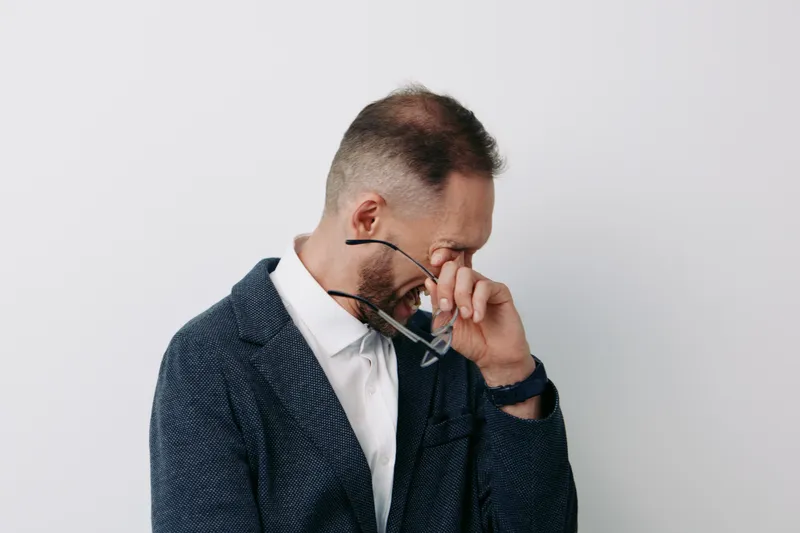 A business professional man in a dark blazer stands against an isolated white background, presenting a composed and focused mood as he adjusts his glasses.