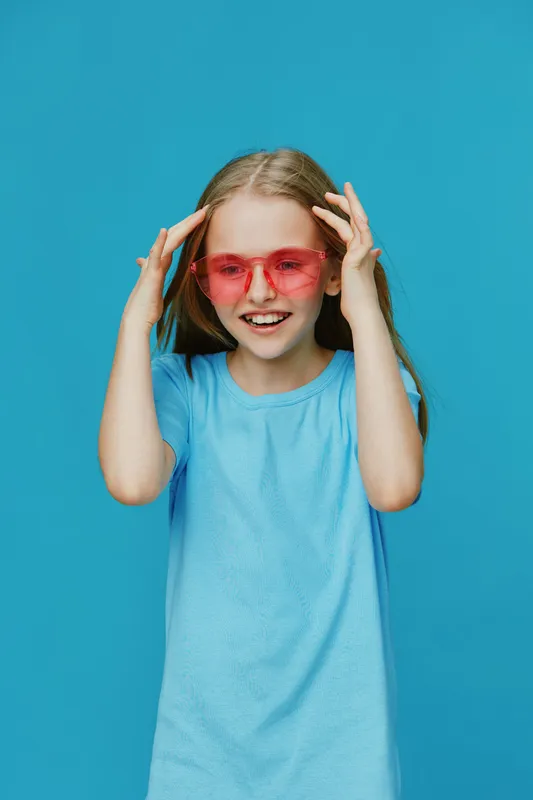 Happy young girl wearing pink sunglasses and a light blue shirt against a vibrant blue background, expressing joy and positivity