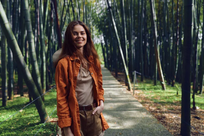 Woman smile bamboo pathway in nature, casual portrait of a young traveler wearing a jacket and relaxed outfit on a scenic forest walkway, candid happy outdoor lifestyle photo