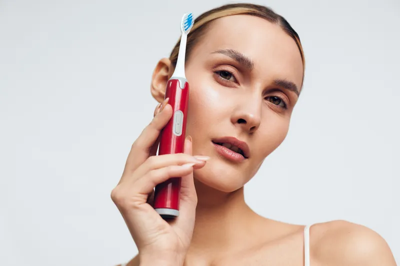 Close-up of a woman holding a red toothbrush near her face, illustrating a daily dental care routine and oral hygiene focus in a bright, clean studio setting with soft lighting.
