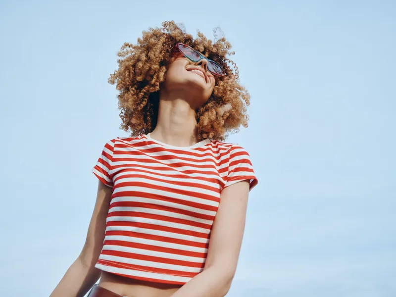 A confident young woman with curly hair stands against a clear blue sky, wearing a red and white striped crop top and sunglasses, conveying carefree summer style.