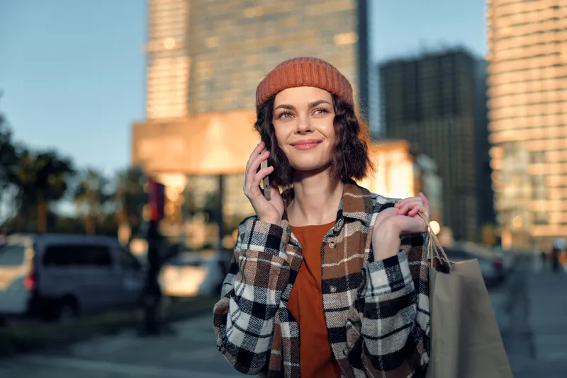 Woman on phone with shopping bag in city street, smile and beanie captured in golden hour glow, candid lifestyle portrait emphasizing authenticity, mindful living and emotional storytelling.