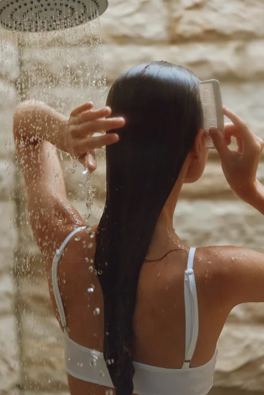 woman combing wet hair under shower with water droplets falling, natural light, smooth skin, relaxed mood, indoor bathroom setting