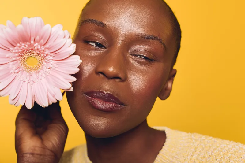 Close-up of a dark skinned African woman holding a pink flower near her face against yellow background. Concept of natural beauty, skincare, wellness, and floral aesthetics with soft lighting.