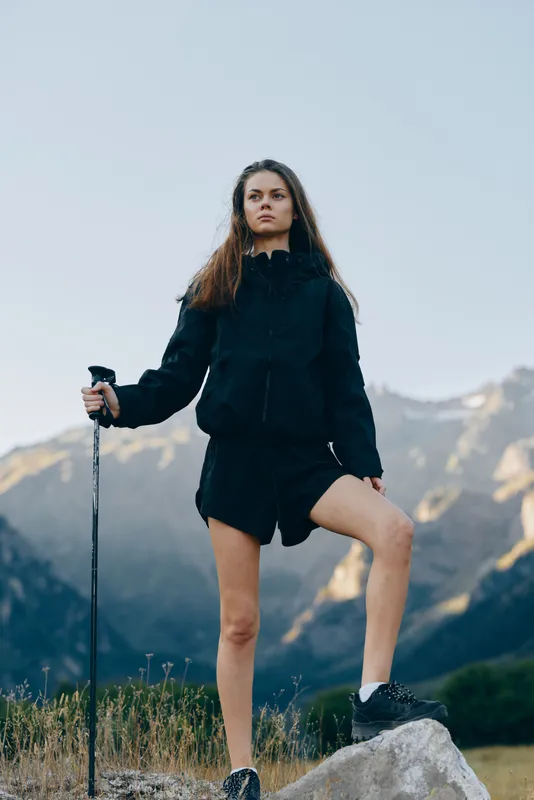 A determined woman stands on a rocky trail with a trekking pole, wearing a dark jacket and shorts. She gazes toward distant mountains, conveying resilience and adventure.