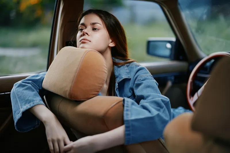 Young woman with long hair resting in a vintage car, exuding a contemplative mood, wearing a denim jacket, surrounded by a natural green background.