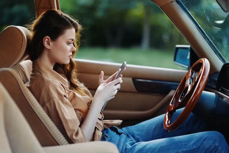 A young woman of Caucasian descent sitting in a vintage car, checking her smartphone, exuding a relaxed mood with soft natural lighting in the background.