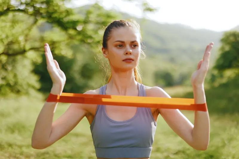 Fit young woman exercising with resistance band outdoors, showcasing strength and determination in a natural green background during summer