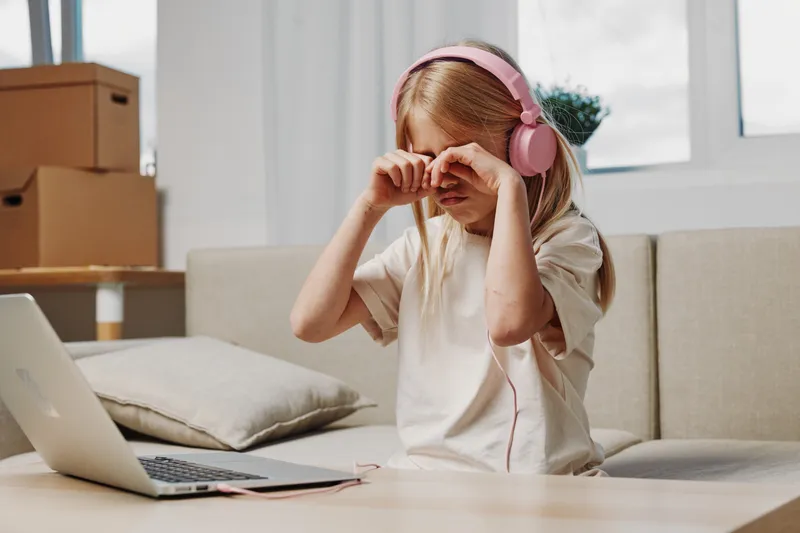Tired child with blond hair wearing pink headphones rubs eyes while sitting on couch with laptop nearby in bright living room.