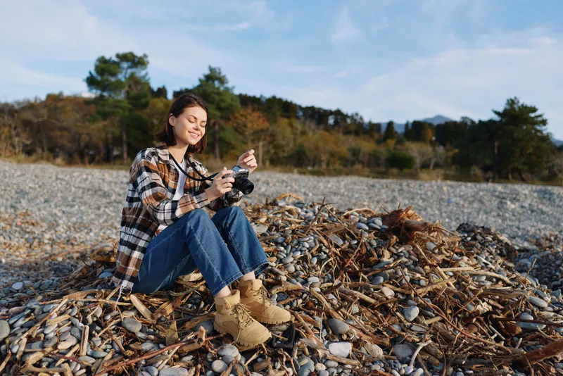 Casual photographer seated on a pebble beach with a camera, wearing a plaid shirt and boots, captured in autumn light among trees and calm shoreline.