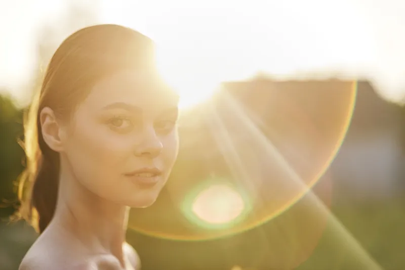 Young woman with glowing sun flare, light skin, natural makeup, outdoor portrait, conveying joy and serenity against a soft golden background