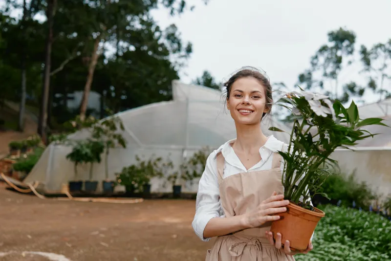 Young woman with a plant in a greenhouse, wearing an apron, showcasing enthusiasm for gardening, surrounded by greenery and natural light