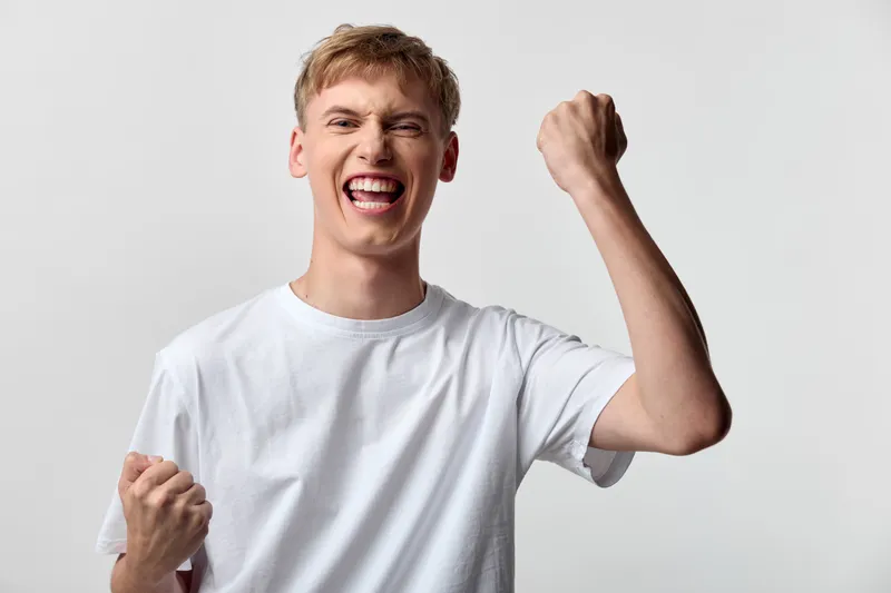 Energetic celebration of victory and joy, a young man pumps fists in triumph, wearing a white shirt, against a bright studio background, with an expressive smile and dynamic feeling of achievement