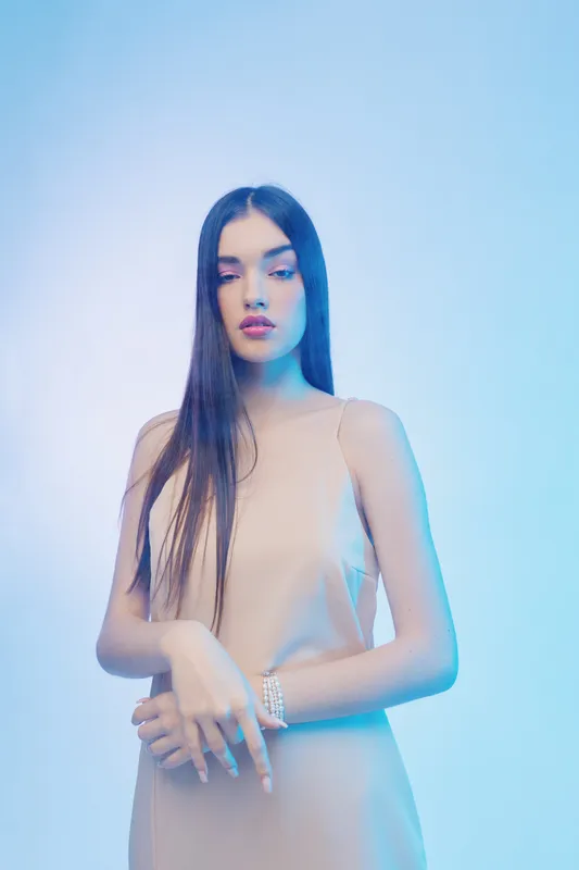 Young woman with long dark hair wearing a beige dress poses with hands crossed in front of a light blue background. Concept of beauty, fashion, and elegance.