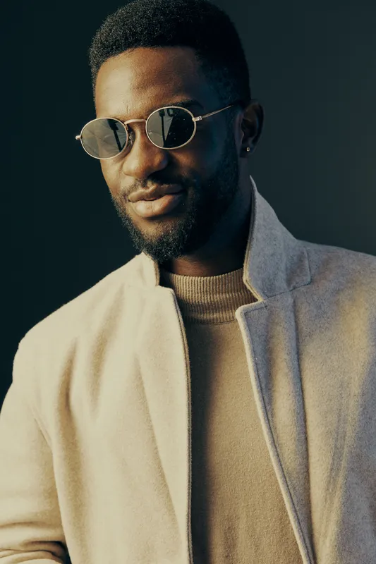 Stylish African man in sunglasses, wearing a light coat and sweater, confidently posing against a dark background, showcasing modern fashion trends