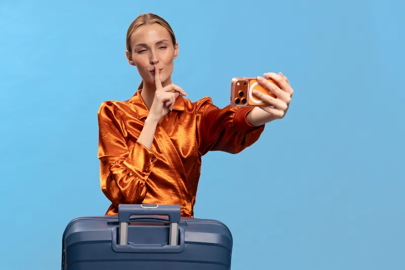 Young woman in orange blouse takes selfie with suitcase, making shushing gesture with finger on lips. Travel, secret, and vacation concept on plain blue background.