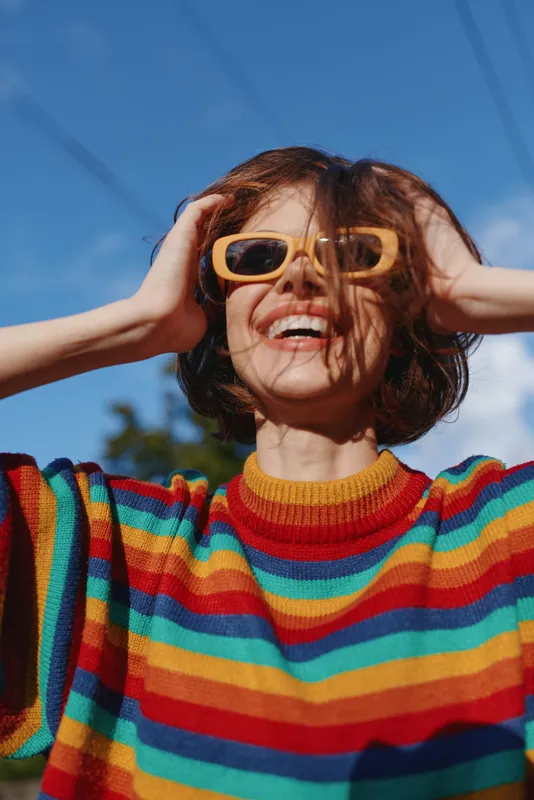 Woman smiling in a colorful striped sweater and yellow sunglasses, short brown hair, joyful millennial with hands on head looking up on a sunny outdoor city street, casual fashion.
