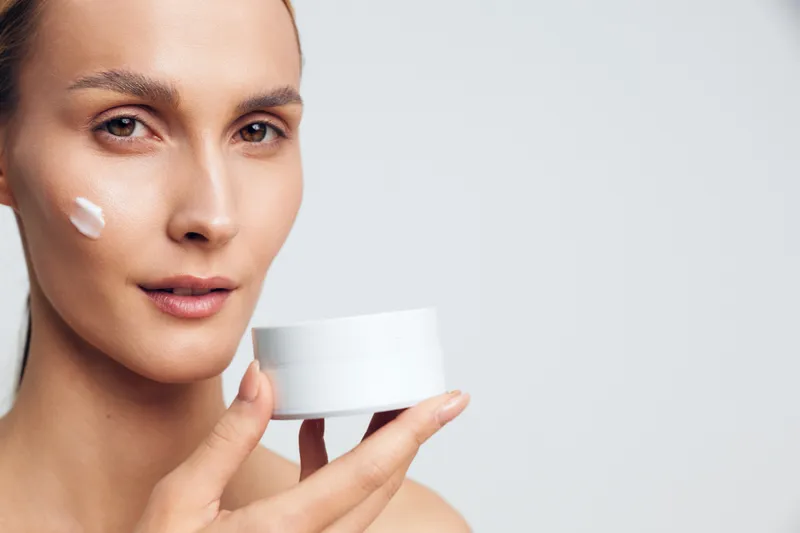 Young woman skincare closeup applying moisturizer with white cream on cheek holding small jar near face clean beauty concept on neutral background spa treatment daily