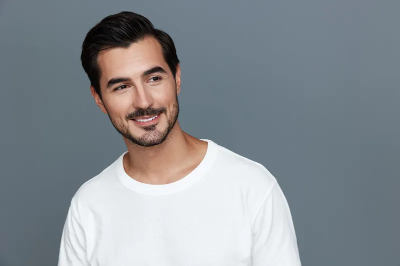 Smiling man wearing a white sweater stands confidently in front of a solid gray background, exuding a warm and inviting demeanor. Lifestyle portrait concept.