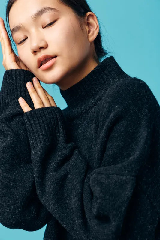 Young woman in black sweater with closed eyes and relaxed expression touching face and neck in soft natural light with blue background for calm and peaceful mood portrait