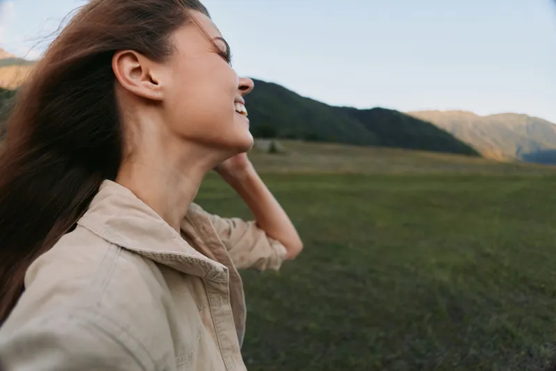 Cheerful woman outdoors, smiling in a sunlit field, relaxed posture, soft breeze tousling hair, natural colors, expansive landscape in the background
