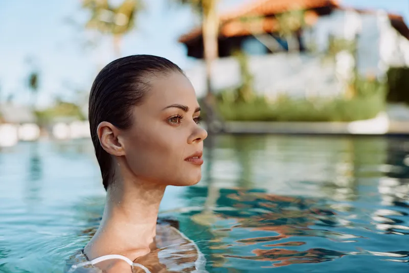 A woman in a swimsuit stands in a sunlit pool, head above water with a calm expression. An outdoor resort backdrop conveys relaxation and serenity in a lifestyle moment.