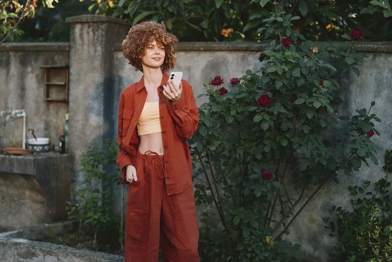 Woman lifestyle portrait outdoors in rust red suit, casual fashion, holding a phone, surrounded by blooming flowers, standing near a garden wall, warm natural light, relaxed mood
