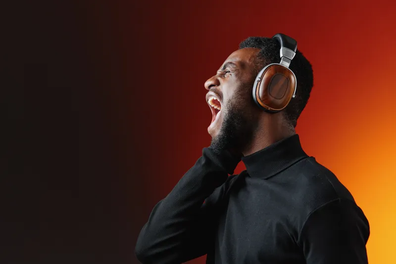 Young Black man enjoying music with headphones, expressing joy and passion, wearing a black turtleneck against a vibrant gradient background