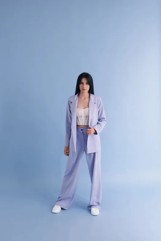 Young woman in pastel lavender suit standing against soft blue background with casual white sneakers, showcasing modern professional fashion style and confident posture.