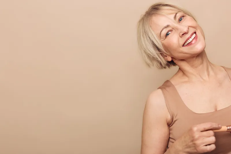 Smiling mature woman with short blonde hair wearing sleeveless top poses against beige background. Concept of happiness, confidence, beauty, and natural aging with soft lighting and warm tones.