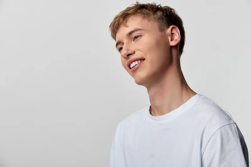 Young man with a bright smile wearing a white tee against a neutral background, showcasing casual style and confident expression suitable for lifestyle, fashion, or wellness imagery
