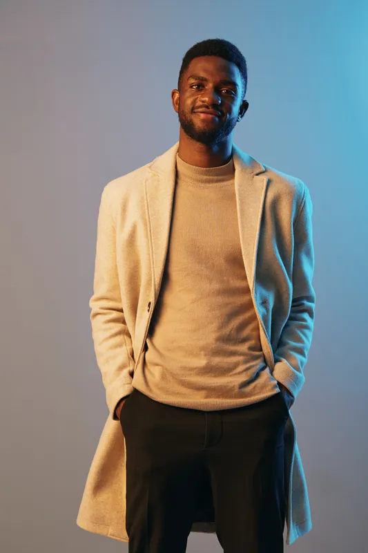 Confident Black man in stylish beige coat and sweater, posing against a blue and gray background with a warm expression, perfect for fashion and lifestyle concepts