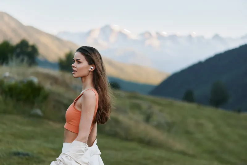 A serene woman stands in a grassy landscape with distant mountains under a clear blue sky. She wears an orange crop top and a light jacket, facing sideways with calm confidence.