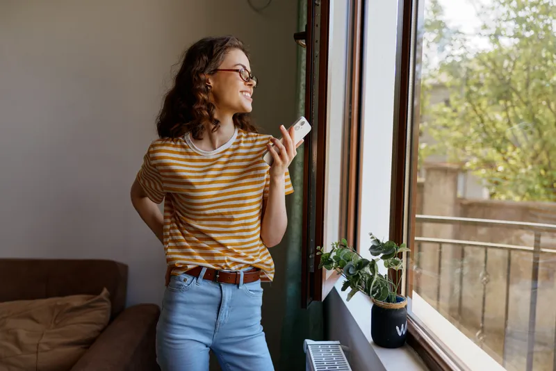 Casual woman indoors by a sunlit window, smiling and relaxed, striped tee and jeans, capturing a simple home lifestyle moment with natural light and greenery nearby and calm atmosphere