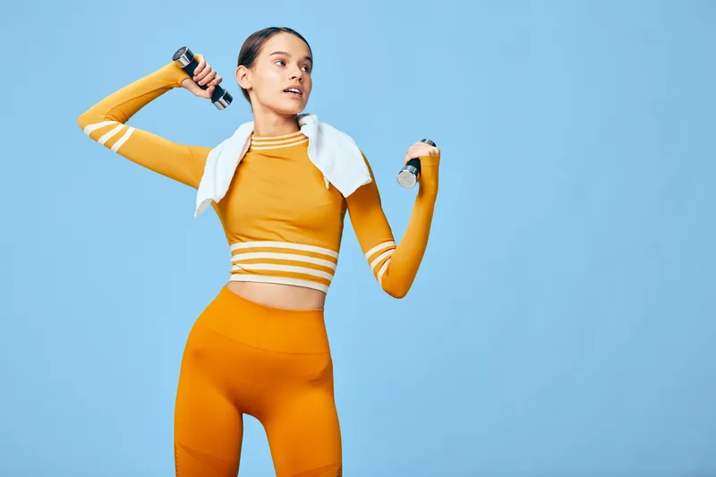 Active woman in bright orange athletic wear lifts dumbbells with a towel over shoulders, mid workout in a vibrant studio, showcasing fitness, strength and energy.