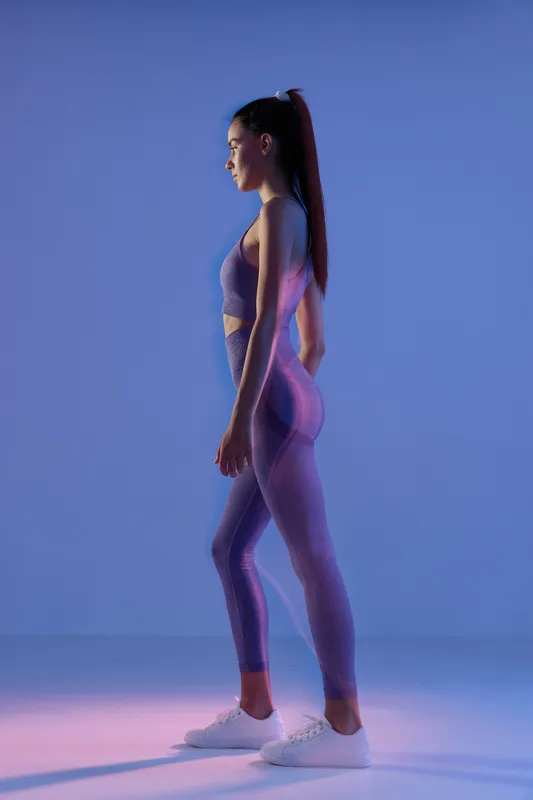 Woman sport fitness wearing purple workout leggings and top stands in profile with focused expression in minimalist blue studio, showcasing active lifestyle and strength.