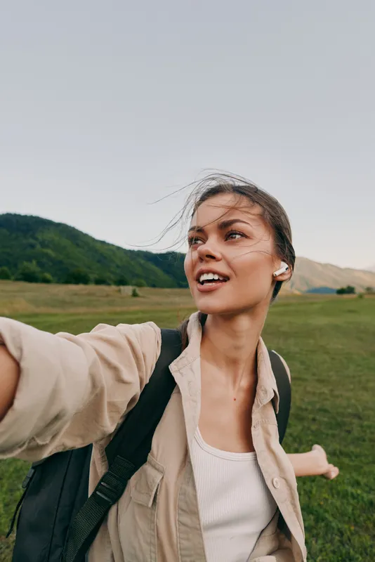 Young woman with backpack taking selfie outdoors in a green field with mountains in the background. Casual lifestyle concept with natural light and fresh air.