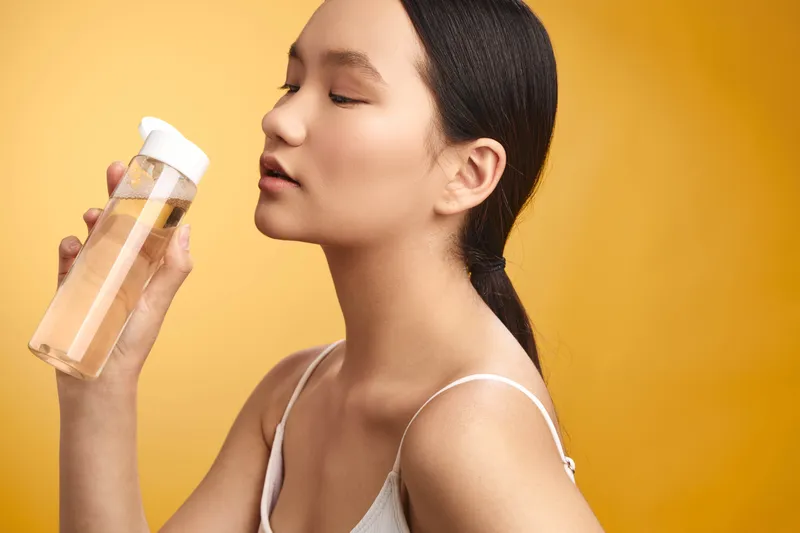 asian woman holds a cosmetic bottle near her face in a bright studio, exuding calm confidence and a fresh skincare vibe that invites care, wellness, and a positive mood.